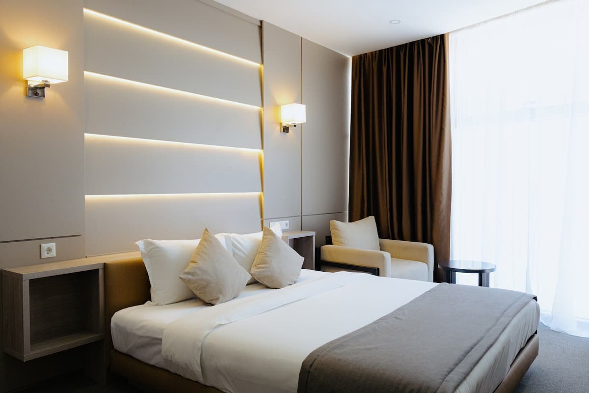 A modern hotel room with a large bed, bedside tables, a chair, and a large window with curtains.