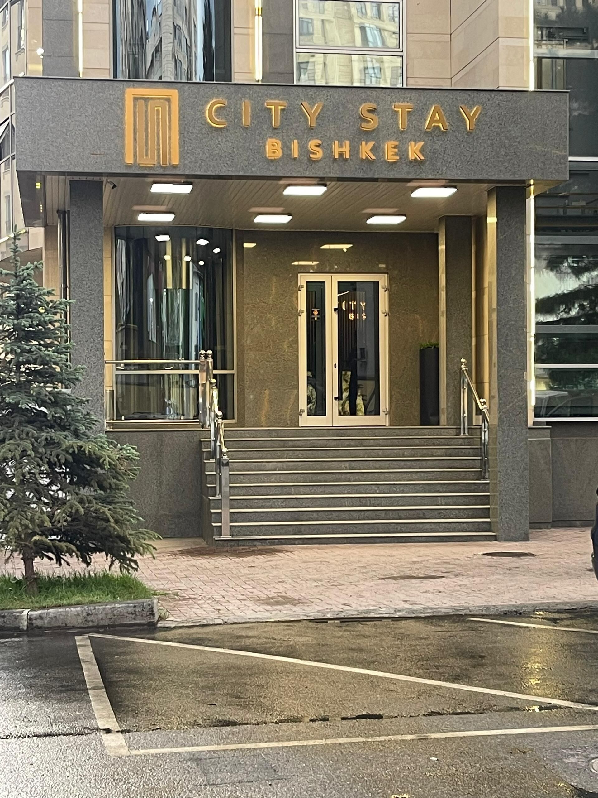 Un immeuble avec une enseigne indiquant « CITY STAY BISHKEK » et un escalier menant à l’entrée.