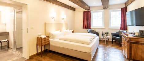 Quarto de hotel com piso de madeira, uma cama grande, uma escrivaninha com cadeira, uma televisão e uma janela com cortinas.