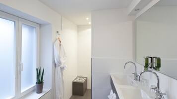 A modern bathroom with a large window, a potted plant, a towel rack, a small stool, and a dual sink with a mirror.