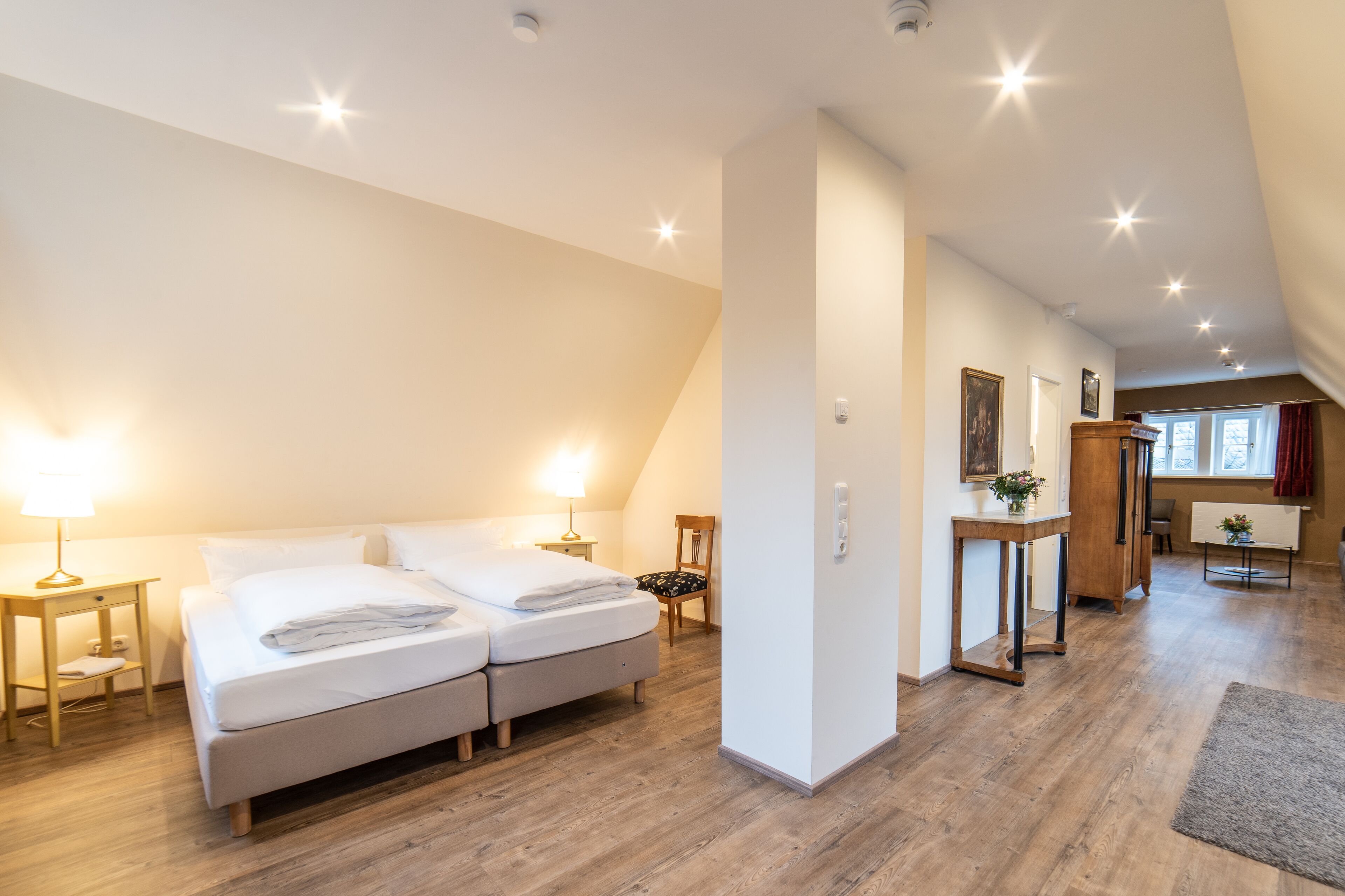 A hotel room with a double bed, a bedside table with a lamp, a wooden floor, and a view of the hallway.