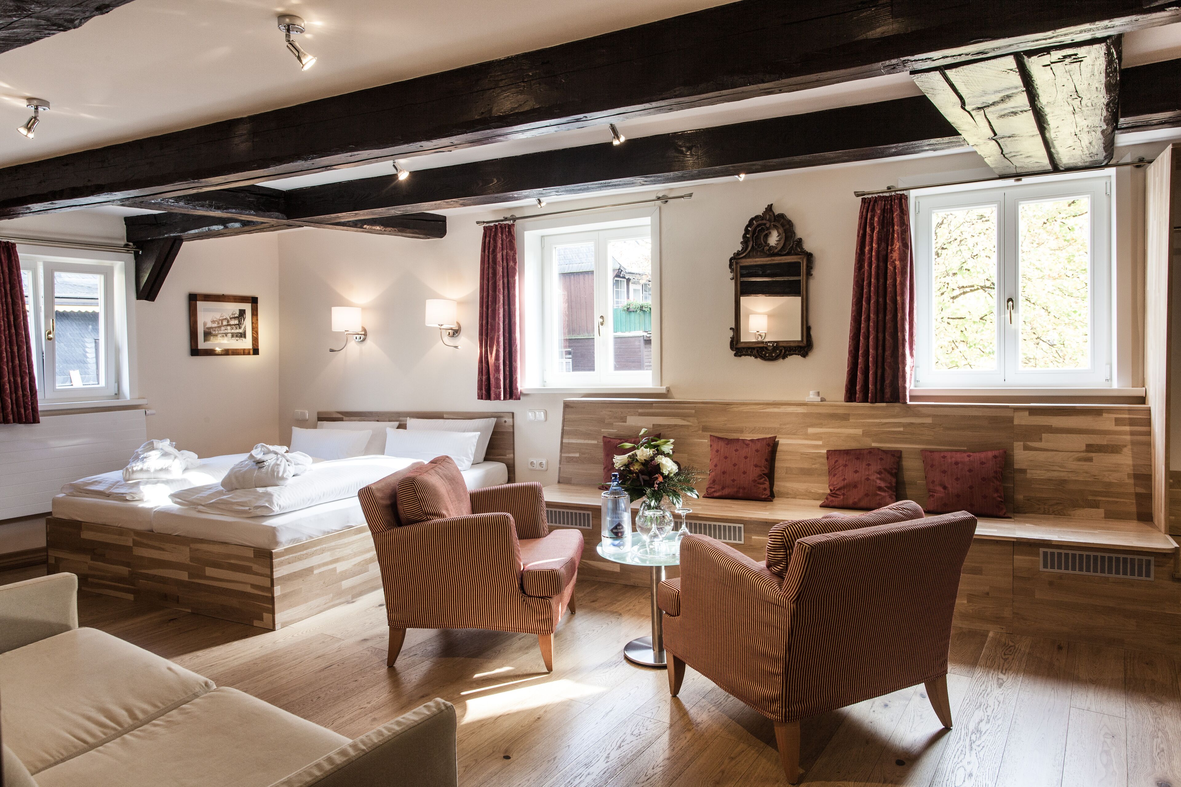 A modern hotel room with a wooden ceiling, a large bed, two armchairs, a sofa, and a small table with a vase of flowers.
