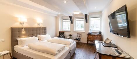 Habitación de hotel con una cama grande, un escritorio con teléfono y papeles, un televisor montado en la pared y dos ventanas con luz natural.