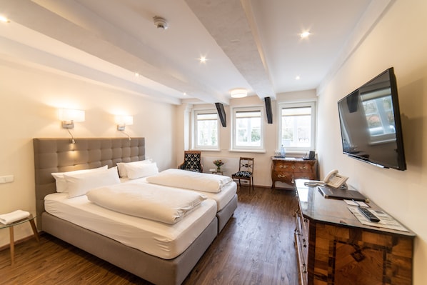 Habitación de hotel con una cama grande, un escritorio con teléfono y papeles, un televisor montado en la pared y dos ventanas con luz natural.