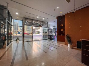 A modern hotel lobby with glass doors leading to a street view, a reception desk, and a potted plant.