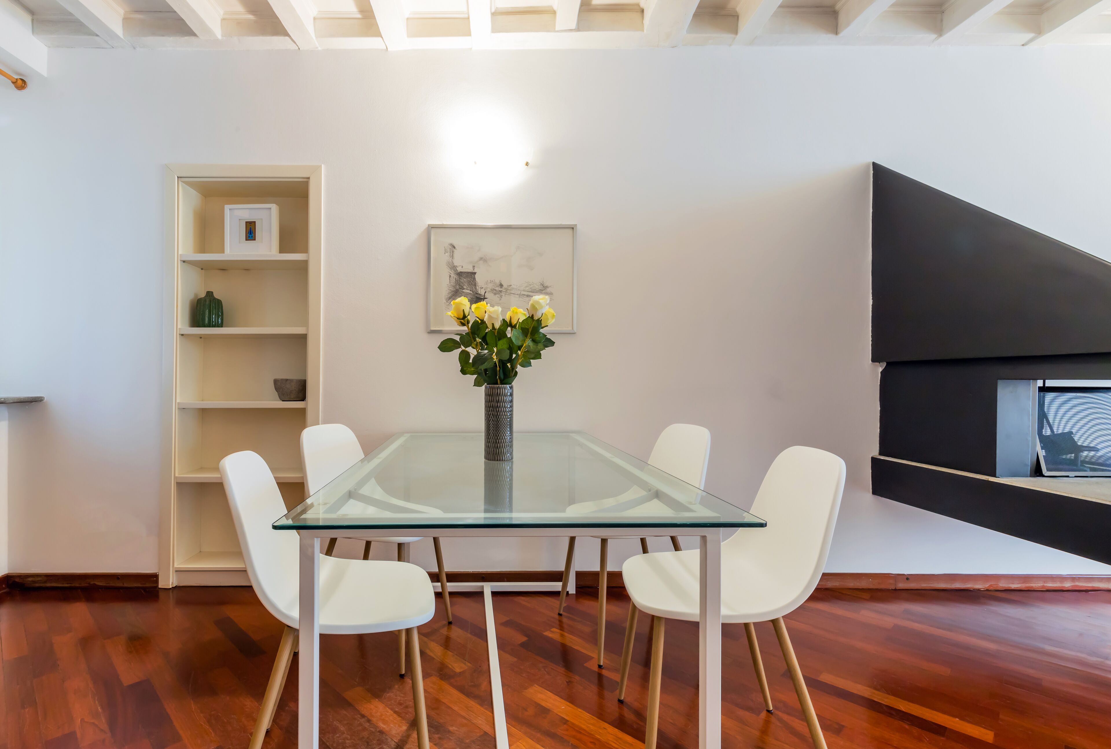 Un comedor moderno con una mesa de vidrio rodeada de sillas blancas, un florero con flores, un estante blanco y una pared de acento negra con un cuadro enmarcado.