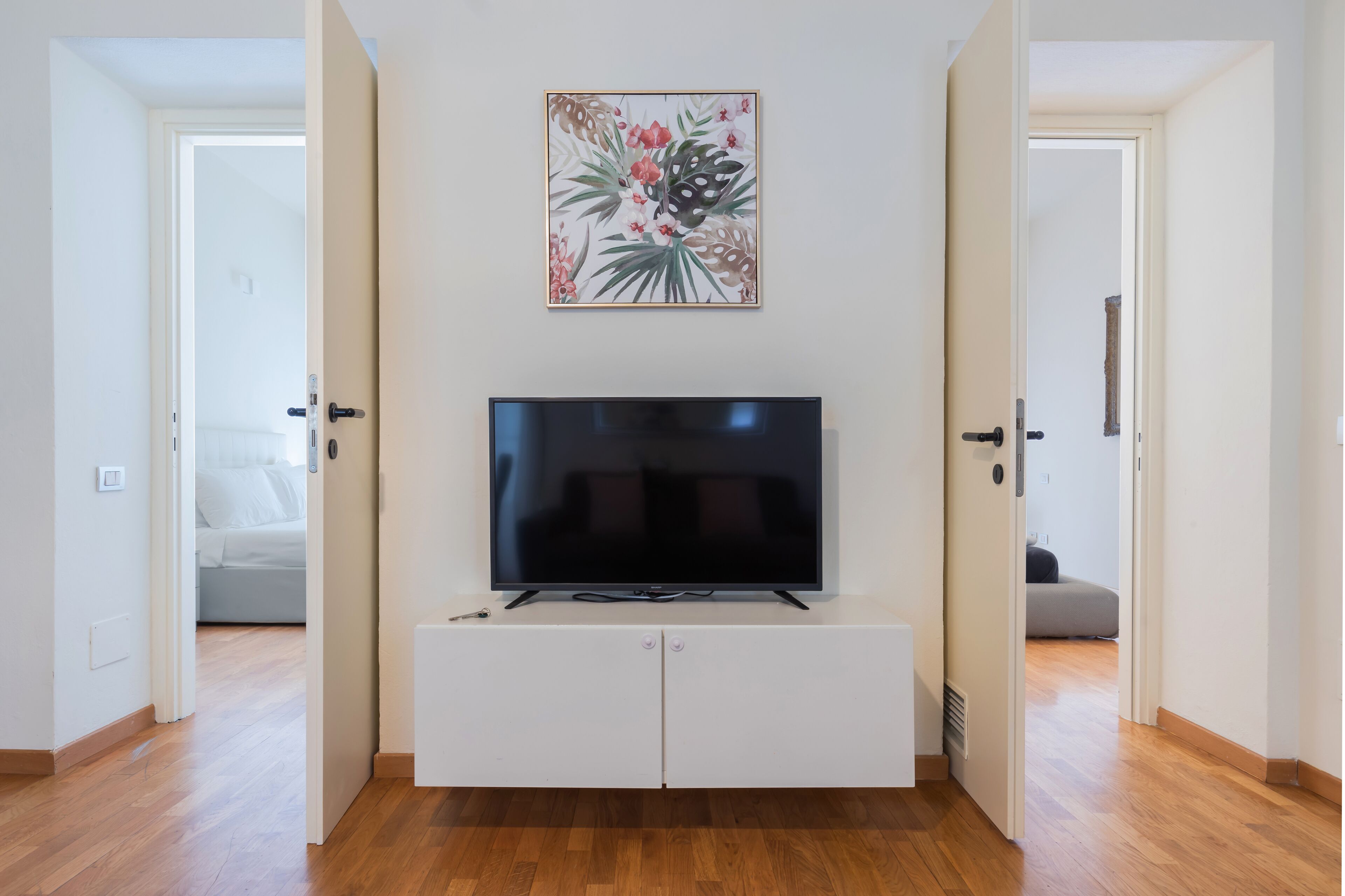 A modern living room with a flat-screen TV on a white cabinet, a floral artwork above the TV, and wooden flooring.