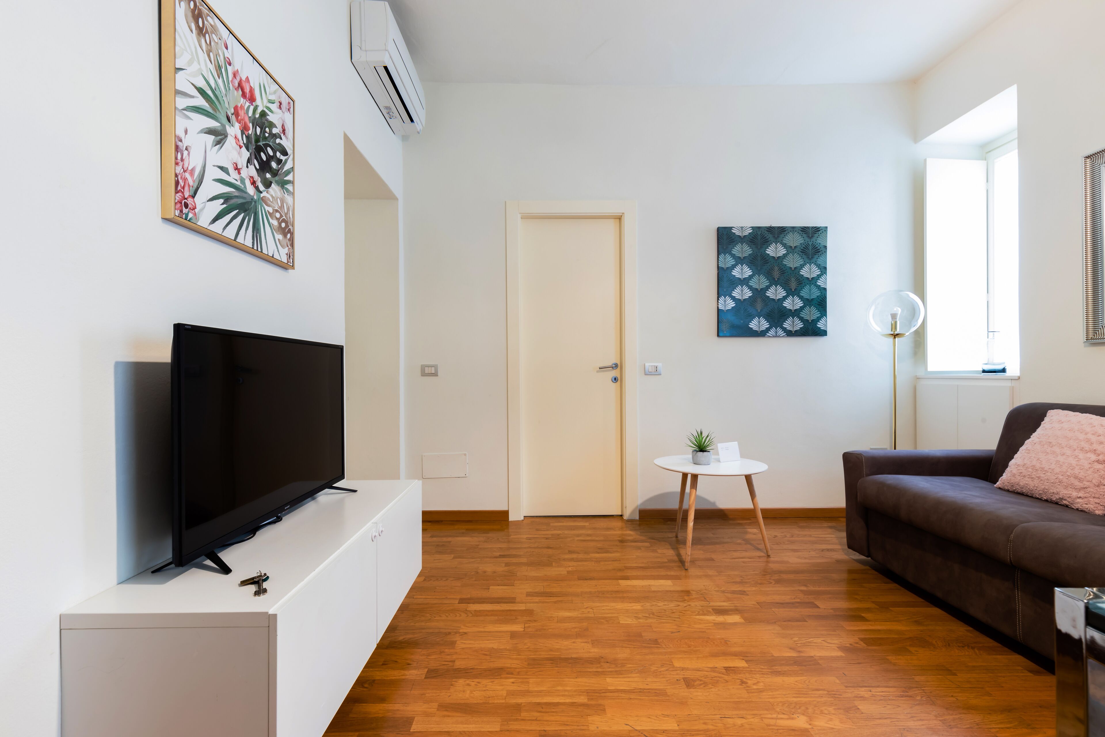 Un salon moderne avec un téléviseur à écran plat posé sur un meuble blanc, un canapé en cuir marron, une petite table ronde agrémentée d’une plante, et une porte donnant sur une autre pièce.