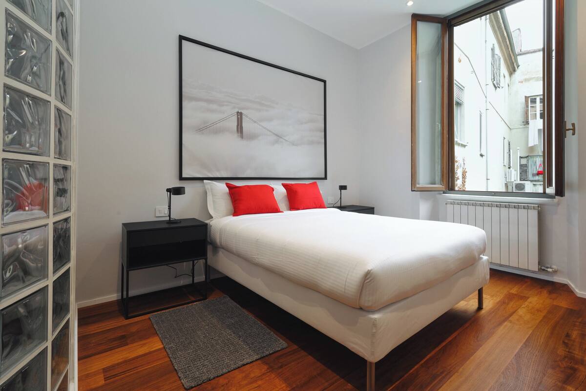 A bedroom with a large bed, a nightstand, a bedside table, a framed picture on the wall, a window with a view of buildings, and a storage unit with clear plastic doors.