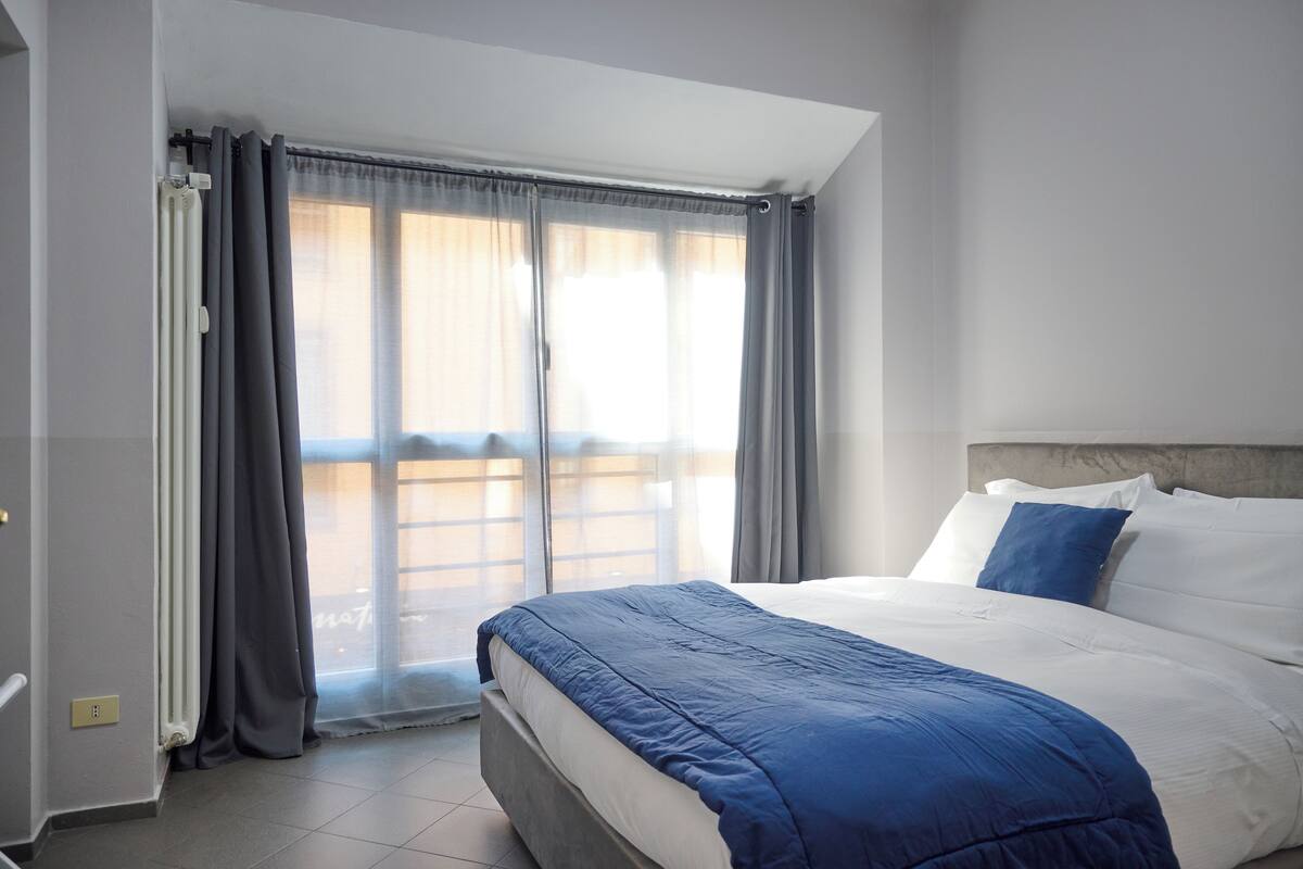 A hotel room with a large bed, a view of the outside through a window with curtains, and a radiator on the wall.