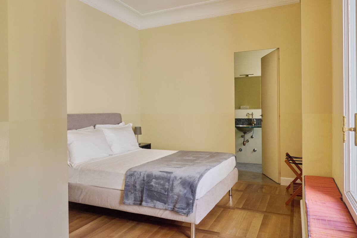 A bedroom with a bed, a nightstand, a mirror, and a bathroom visible through an open door.