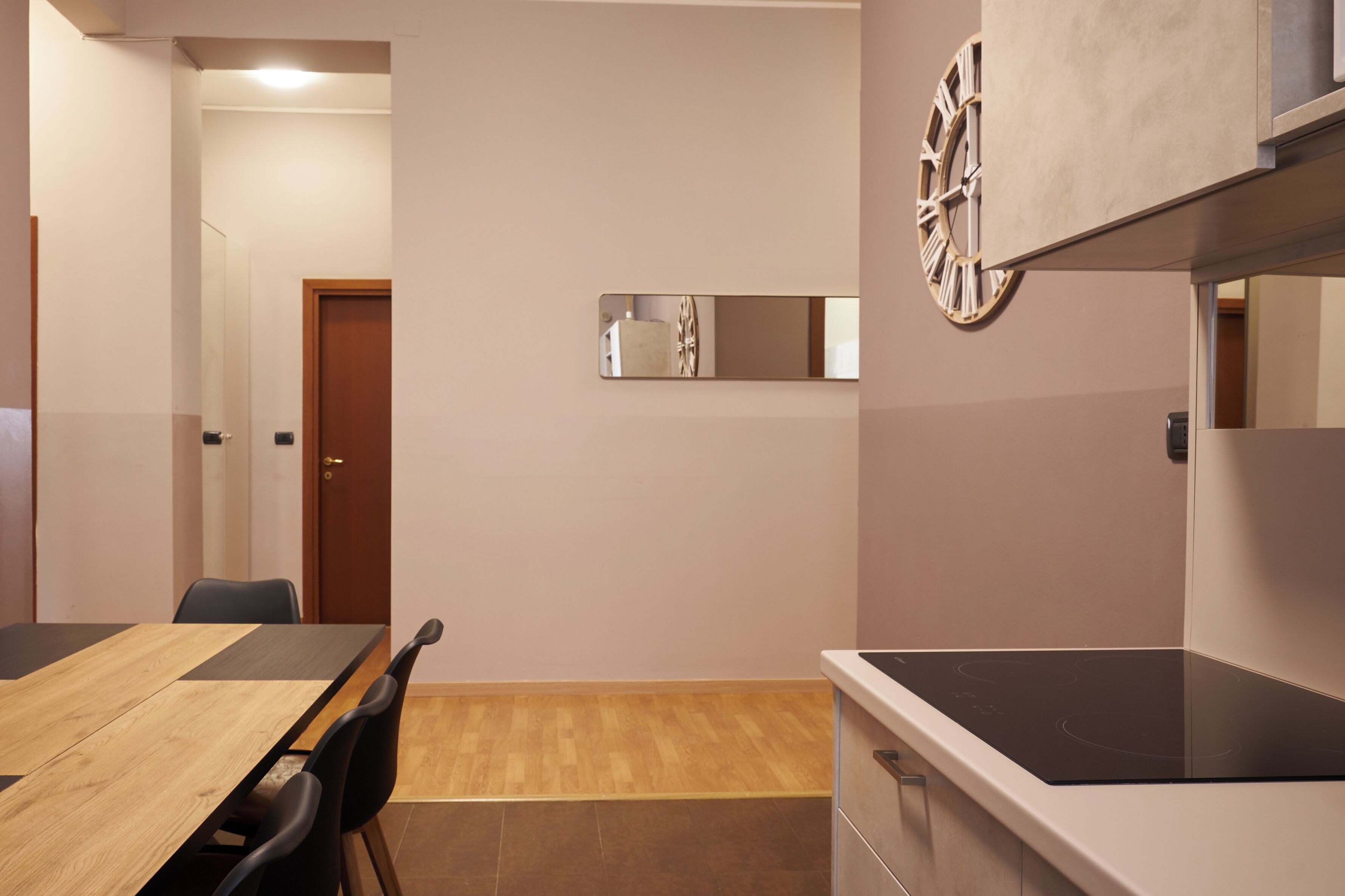 Un espace repas moderne avec une table et des chaises en bois, une cuisine dotée d’une plaque de cuisson à induction noire et une horloge murale.