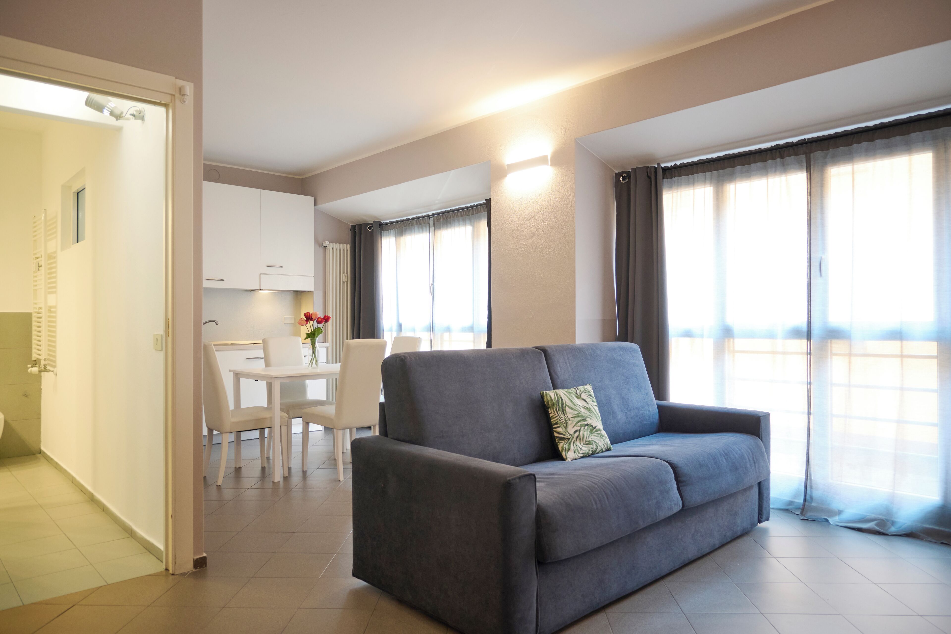 Ein modernes Wohnzimmer mit grauem Sofa, einem Essbereich mit weißen Stühlen und einer Küche im Hintergrund.