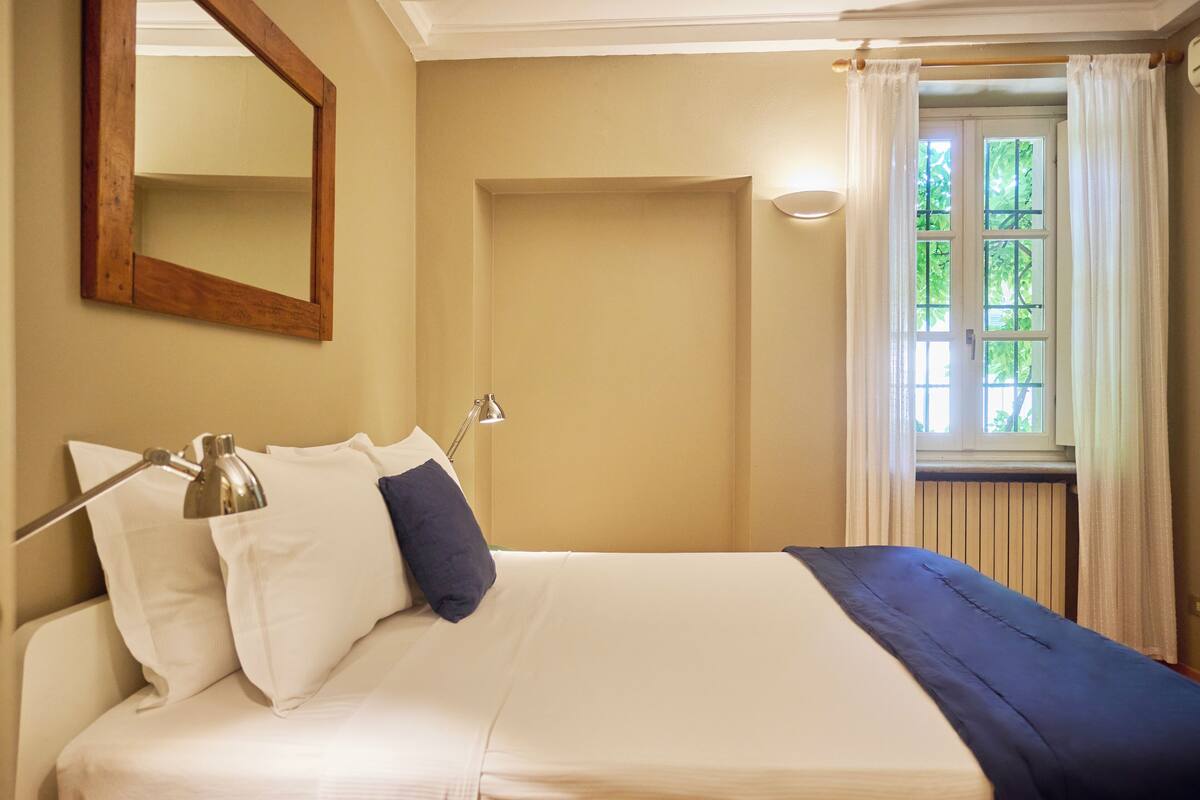 A bedroom with a bed, a bedside lamp, a window with curtains, and a mirror on the wall.
