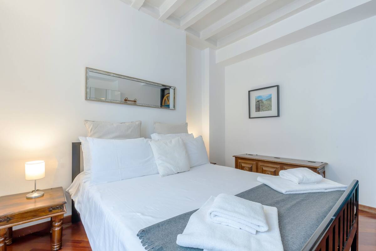 A neatly made bed with white linens, a bedside table with a lamp, a wooden nightstand, a dresser, and a framed picture on the wall.