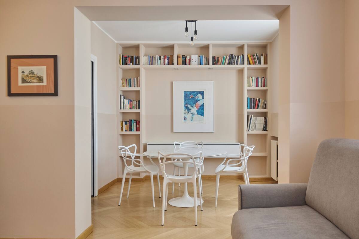 A room with a white dining table, white chairs, and a grey sofa. There are bookshelves with books and a framed picture on the wall.