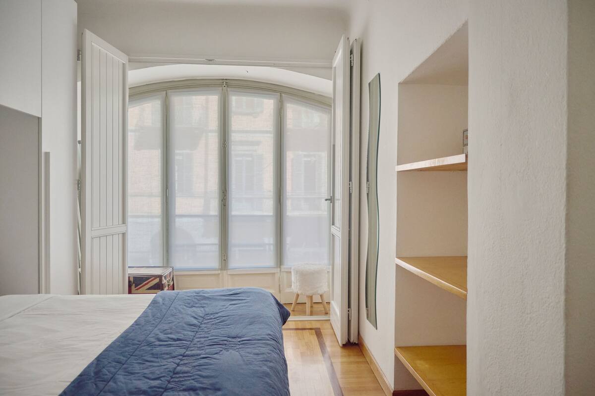 A bedroom with a bed, a wardrobe, a small table, and a window with a view of buildings.