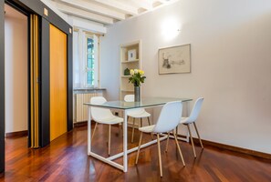 Ein zeitgemäßer Essbereich mit einem Glastisch, weißen Stühlen, einer Blumenvase und einem gerahmten Kunstwerk an der Wand.