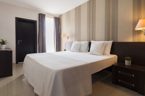 A hotel room with a large bed, a bedside table with a lamp, a dark nightstand, and a potted plant. - Valletta view Apartments by ST Hotels (Gzira)