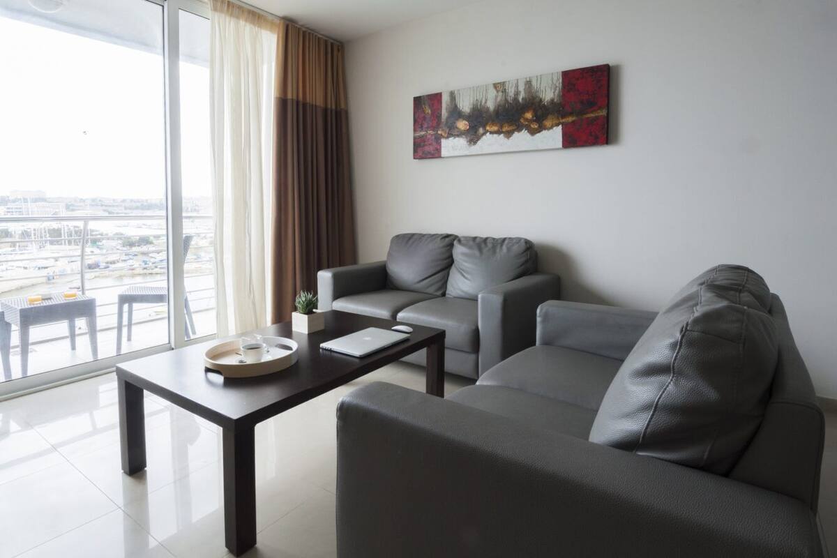 Una sala moderna con un sofá, una mesita de centro y un ordenador portátil. Se ve una ventana con cortinas y vistas a la ciudad.