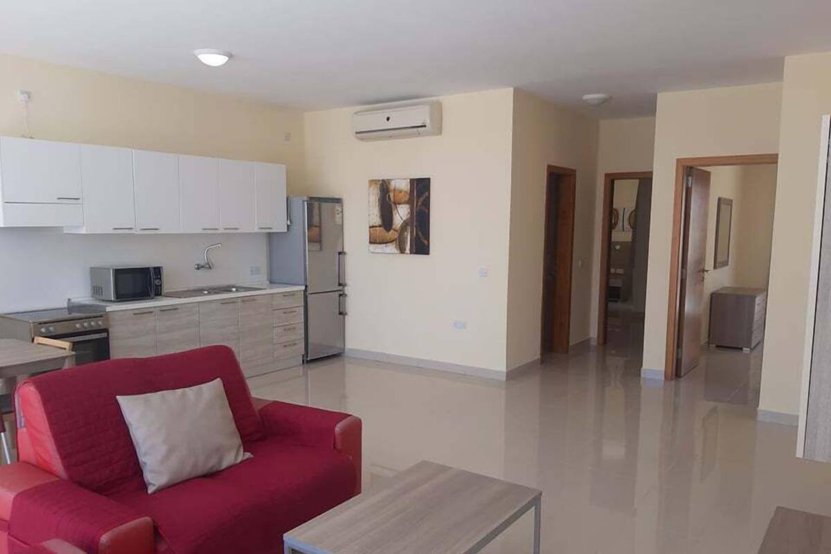 Un apartamento moderno con un sofá rojo, una pequeña mesa de madera, una cocina con armarios blancos y un refrigerador.