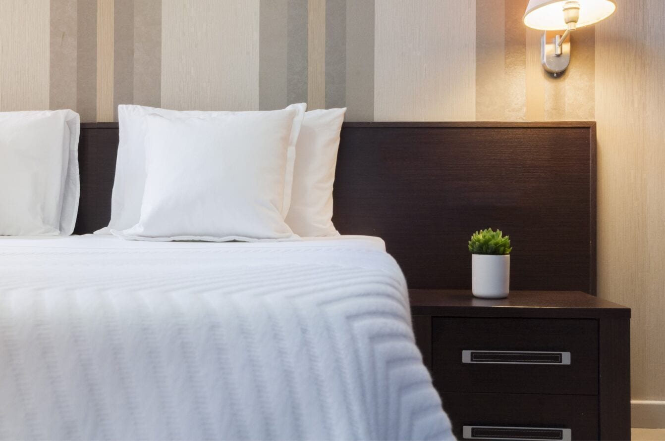 Una cama bien hecha con sábanas y almohadas blancas, una mesita de noche con una planta y una lámpara de pared.