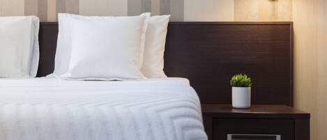 Una cama bien hecha con sábanas y almohadas blancas, una mesita de noche con una planta y una lámpara de pared.