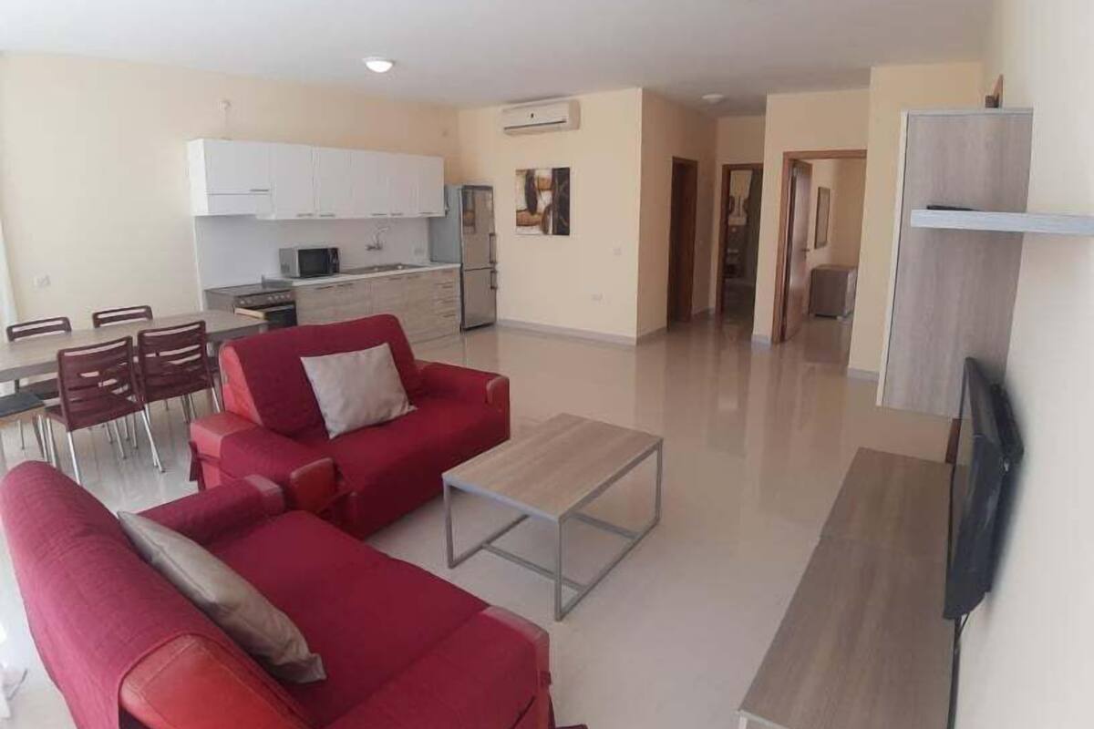 Un apartamento moderno con una sala de estar que presenta un juego de sofá rojo, una mesa de centro y un televisor de pantalla plana. La cocina tiene armarios blancos y un comedor con mesa y sillas.