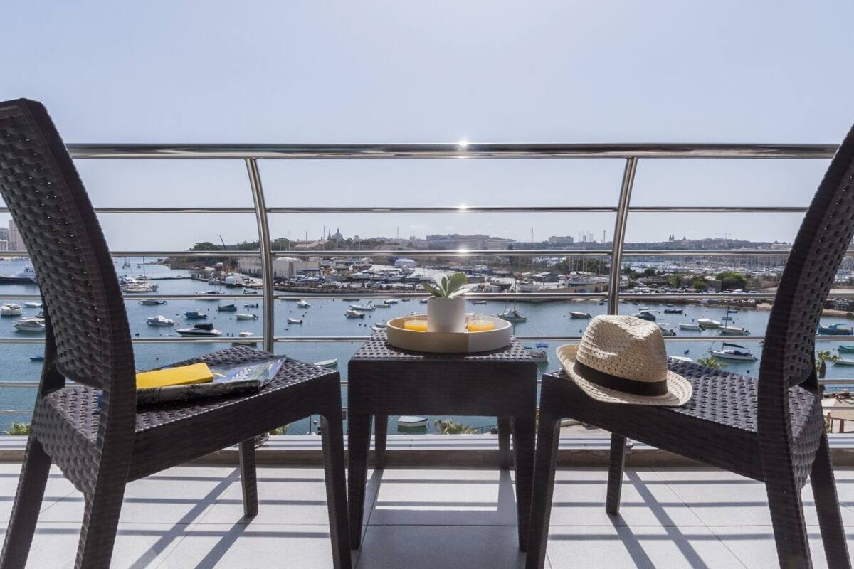 Un balcón con sillas de mimbre, una mesita con un sombrero y vista a un puerto deportivo con barcos y un puente al fondo.
