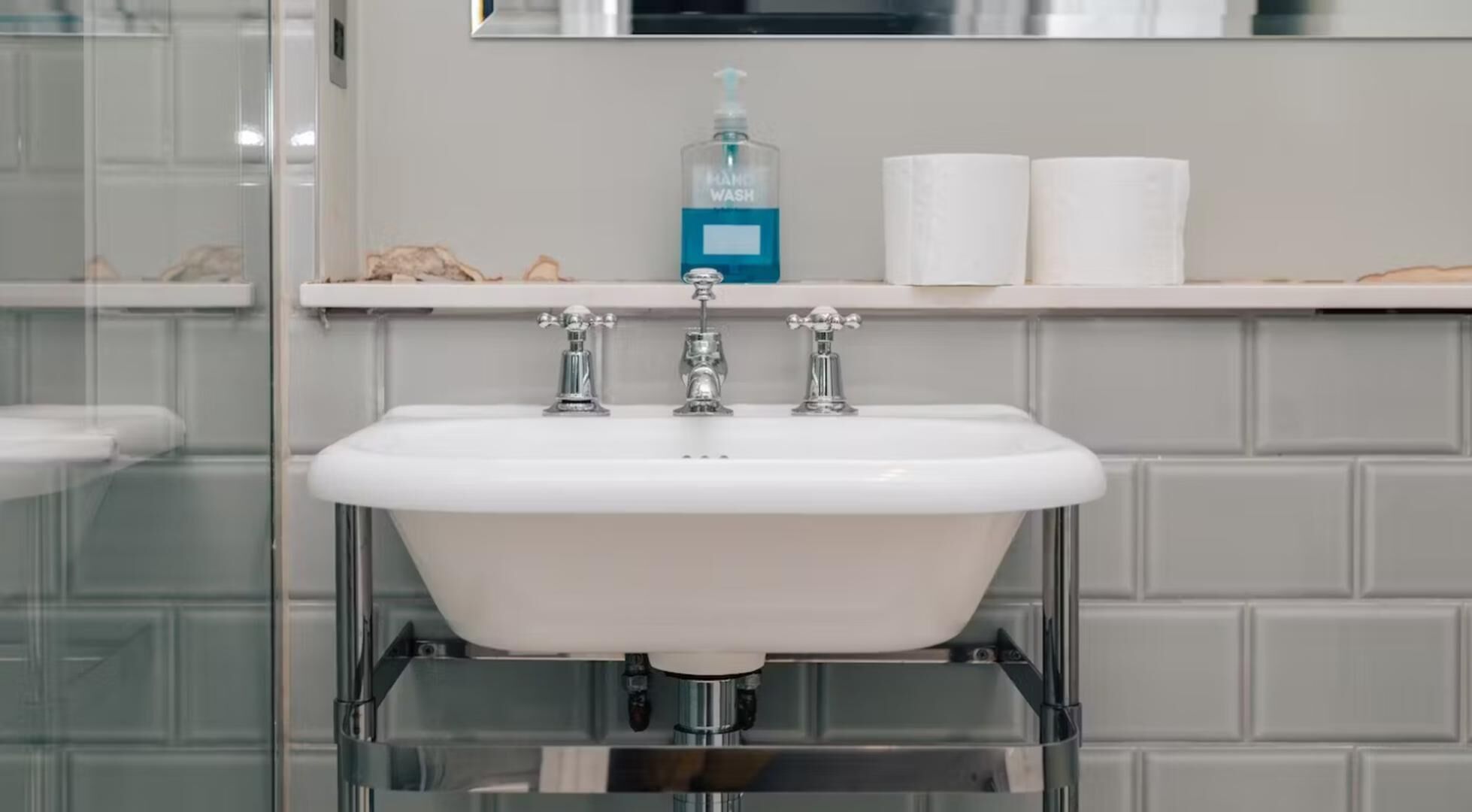 A bathroom with a white sink, three faucets, a soap dispenser, and two rolls of toilet paper on a shelf.