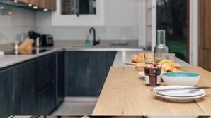 A modern kitchen with dark cabinetry, a wooden countertop, and a breakfast setting including croissants, a bowl of cereal, and a jar of jam.