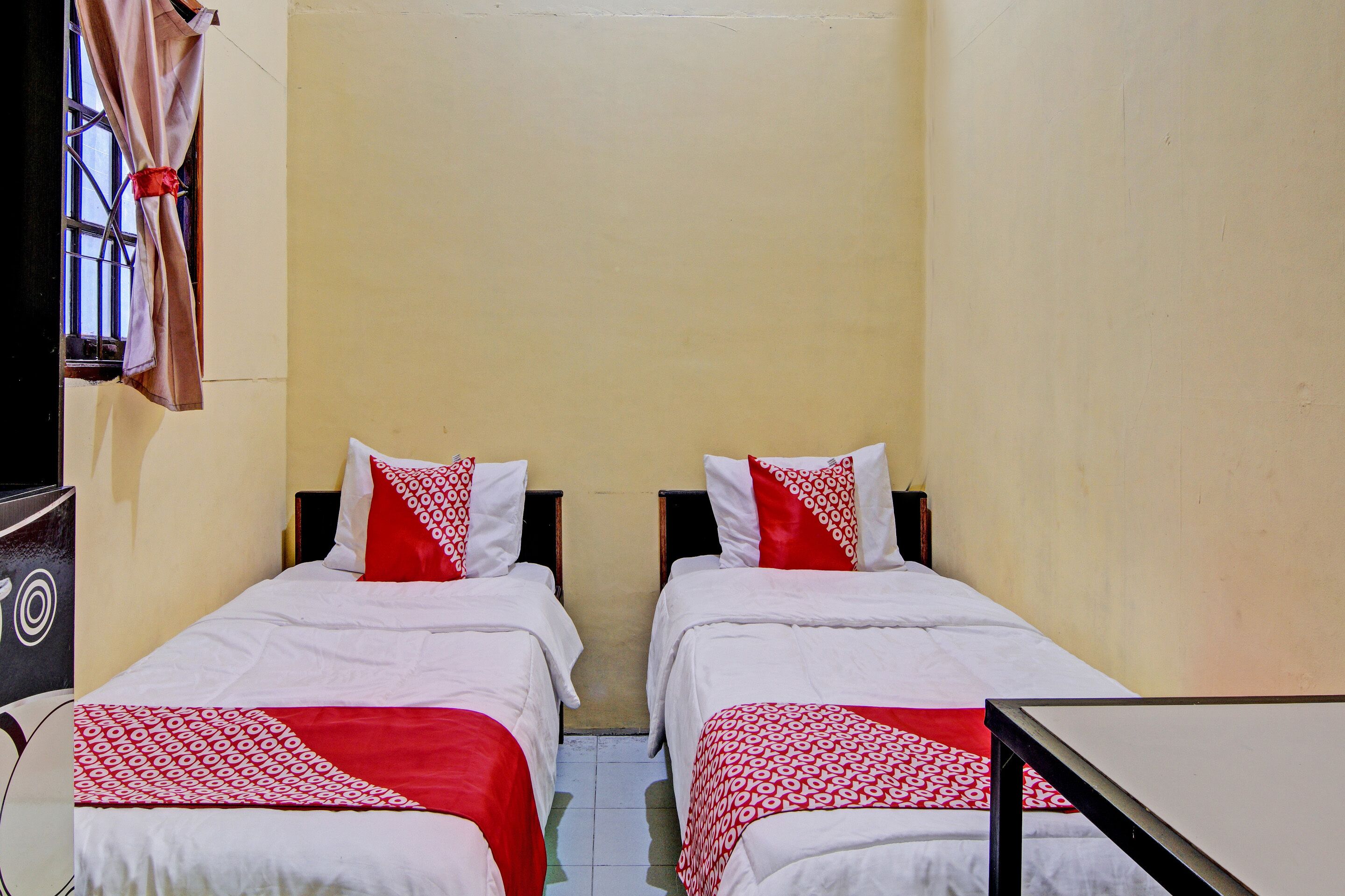 Une petite chambre, de taille modeste, avec deux lits simples, chacun recouvert d’une courtepointe aux motifs rouges et blancs, assortie à des coussins. Les murs de la chambre sont de couleur jaune unie et il y a une fenêtre avec un rideau.