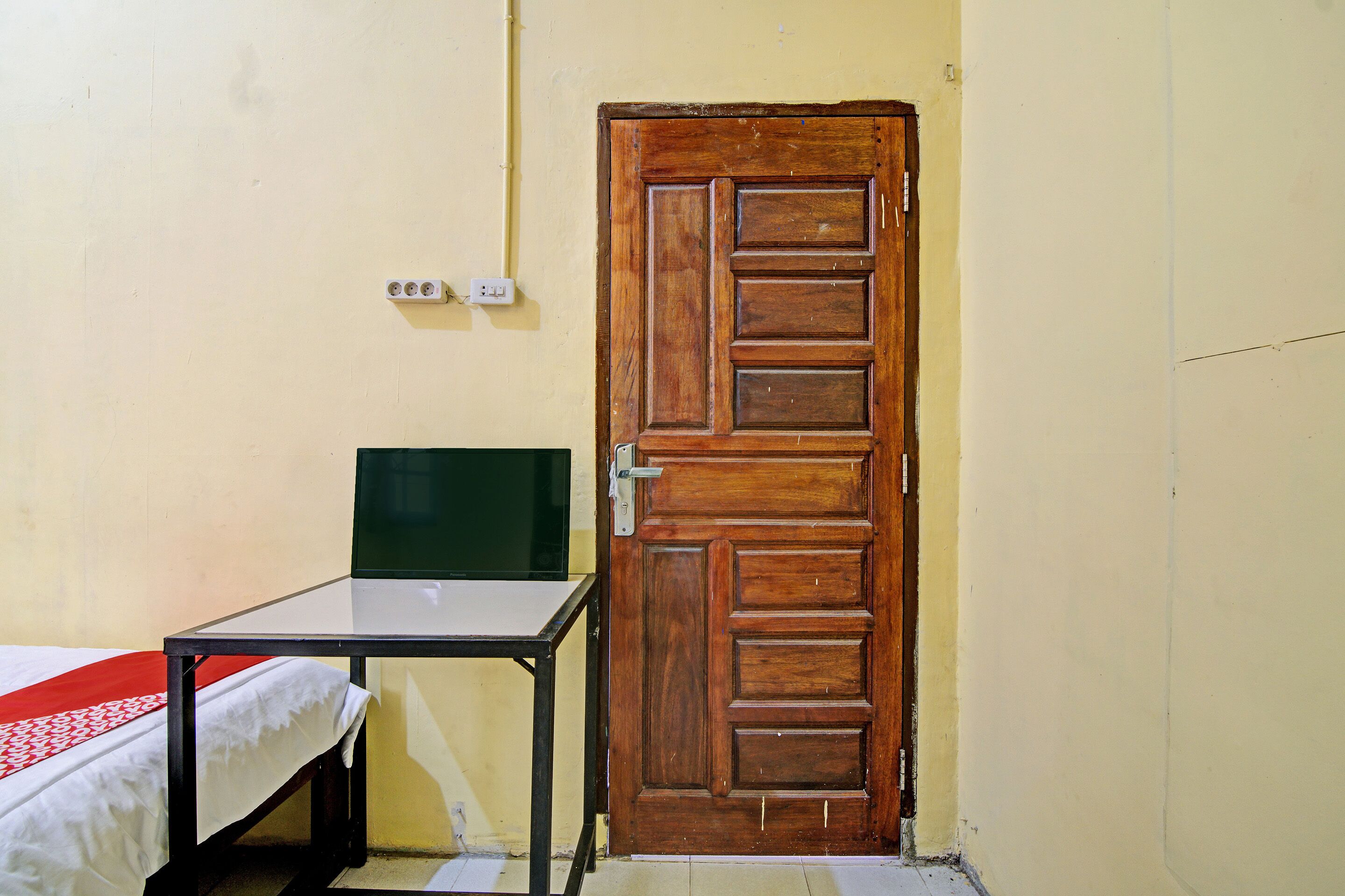 A small room with a wooden door, a bed with a white and red bedspread, and a small table with a green object on it.