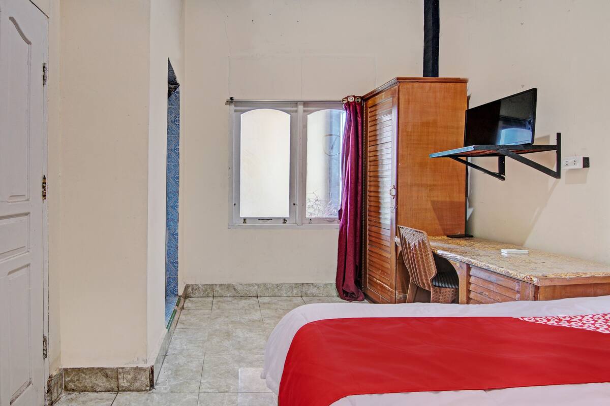 A bedroom with a bed, a desk, a chair, a television, and a window.