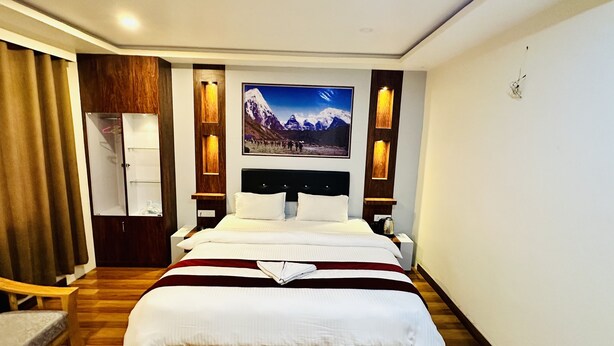 A hotel room with a large bed, wooden headboard, a mounted picture of a mountain landscape, and a wooden wardrobe. - Hotel Pyarij Taplejung (Taplejung)