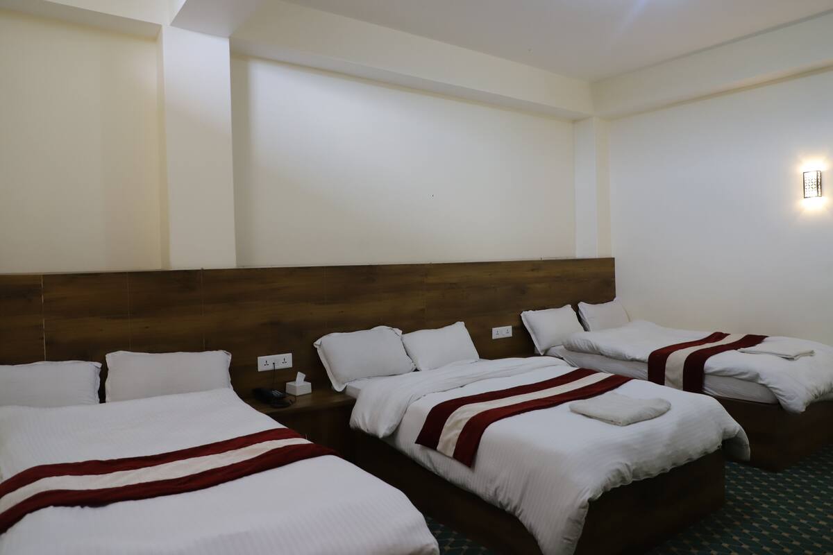 A hotel room with three beds, each with white bedding and red accents, a wooden headboard, and a small bedside table with a lamp.