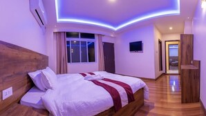A modern bedroom with a wooden headboard, a large bed with white bedding and a red accent, a window with curtains, and a hallway leading to another room. - Hotel Pyarij Taplejung (Taplejung)