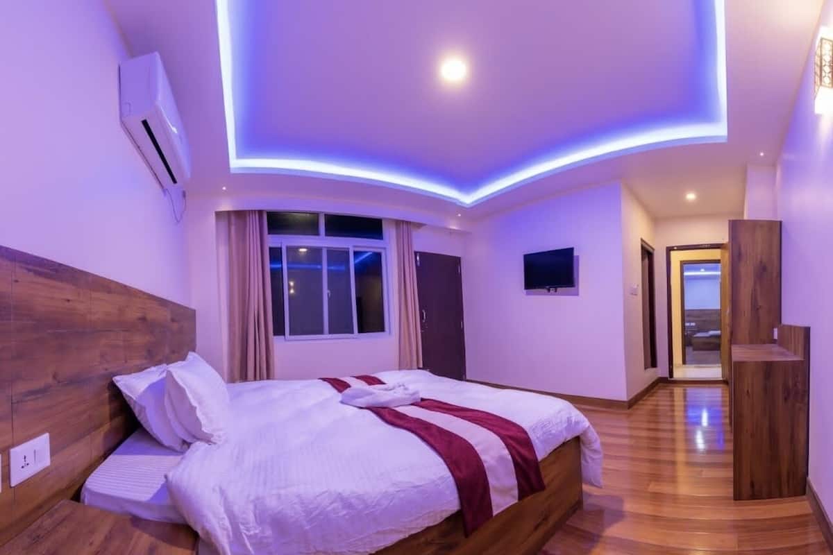 A modern bedroom with a wooden headboard, a large bed with white bedding and a red accent, a window with curtains, and a hallway leading to another room.