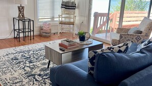 A living room with a blue sofa, a coffee table, a TV, and a view of the outdoors. - GOTL Strip - Private Beach - Lake Erie Views- Sunset Vacation Rentals (Geneva)