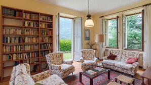 Uma sala de estar aconchegante com uma grande estante de livros, sofás com estampa floral e vista para o verde pelas janelas.
