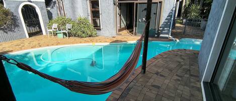 A swimming pool with a hammock, a brick patio, and a building with a unique architectural style.