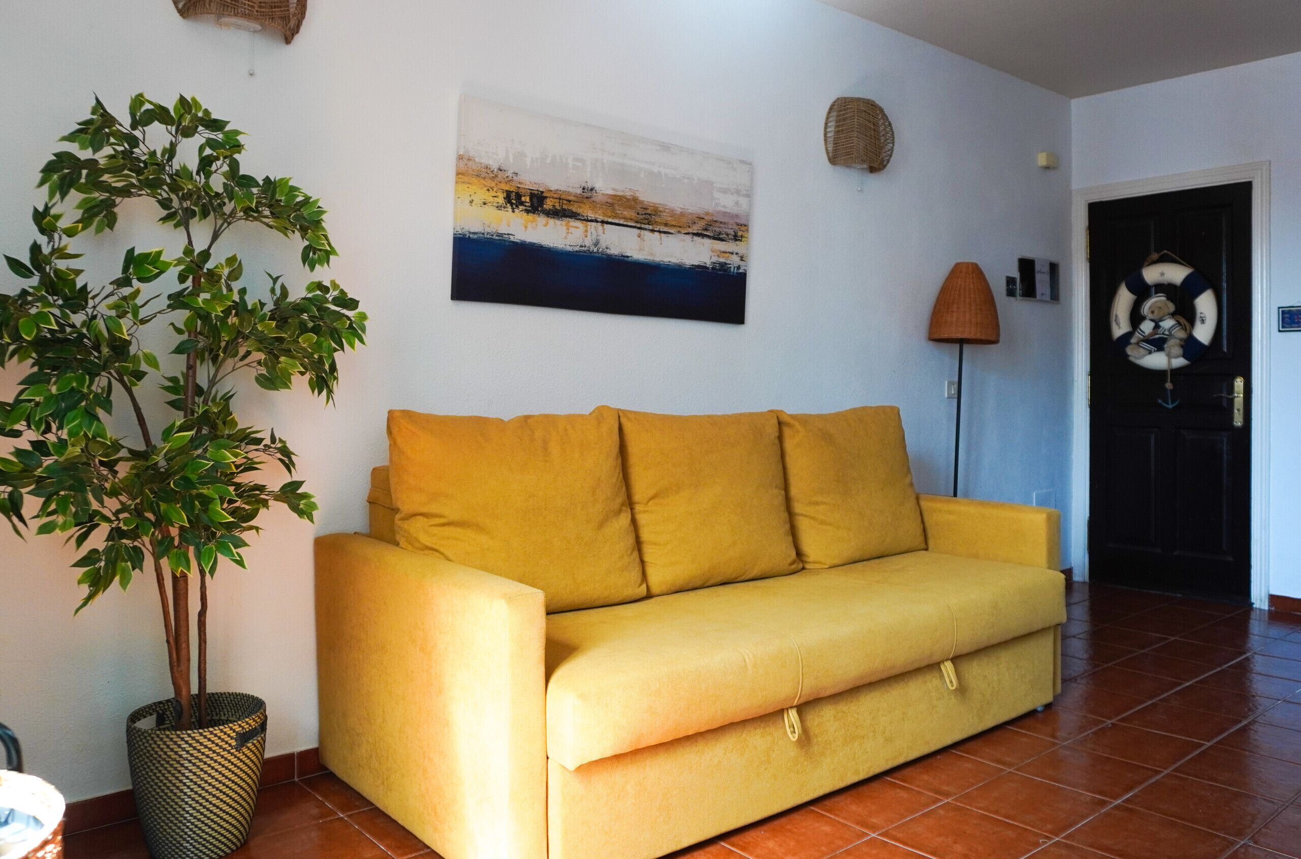 A yellow sofa, a potted plant, a wall-mounted abstract painting, a lamp, and a door with a life preserver decoration.