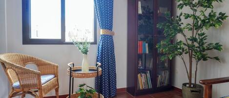 A room with a wicker chair, a small table with a vase and plant, a bookshelf, and a potted tree.