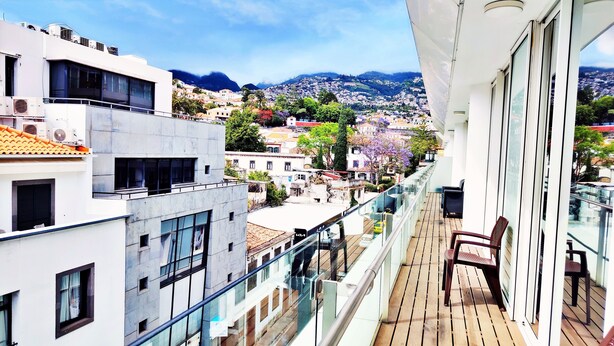 A balcony with a view of a hillside town, a chair, and a glass railing. - Dorisol IV - Luxury Apartment in La Vie (Funchal)