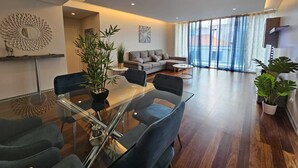 A modern living room with a glass dining table, dark sofas, and a large window with sheer curtains. - Dorisol IV - Luxury Apartment in La Vie (Funchal)