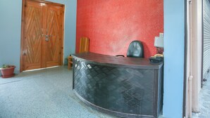A reception area with a wooden door, a red textured wall, a curved reception desk, a chair, and a potted plant. - Hotel Pine & Dine Almora (Almora)