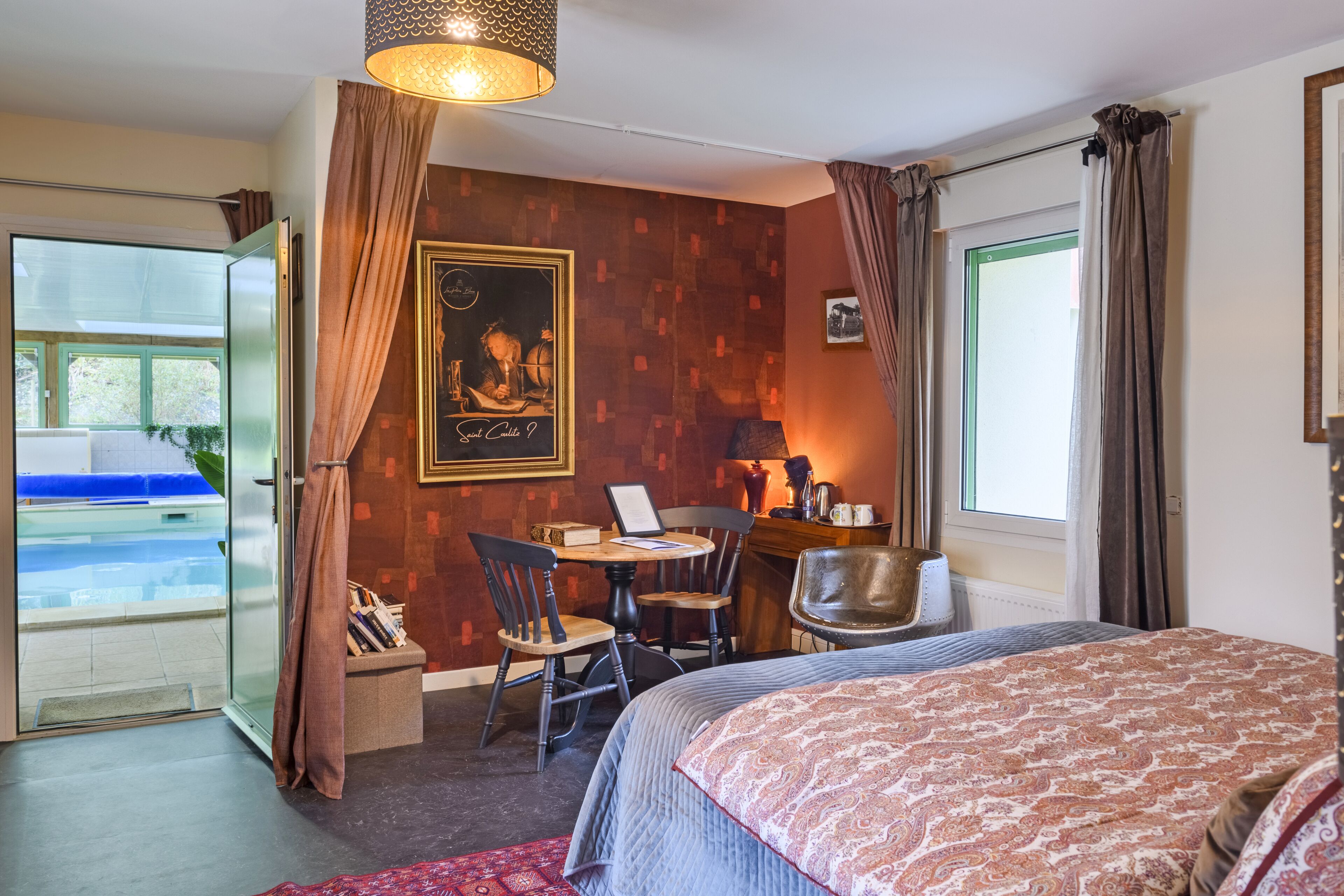 A hotel room with a bed, a desk with a chair, a small table, and a window with curtains.