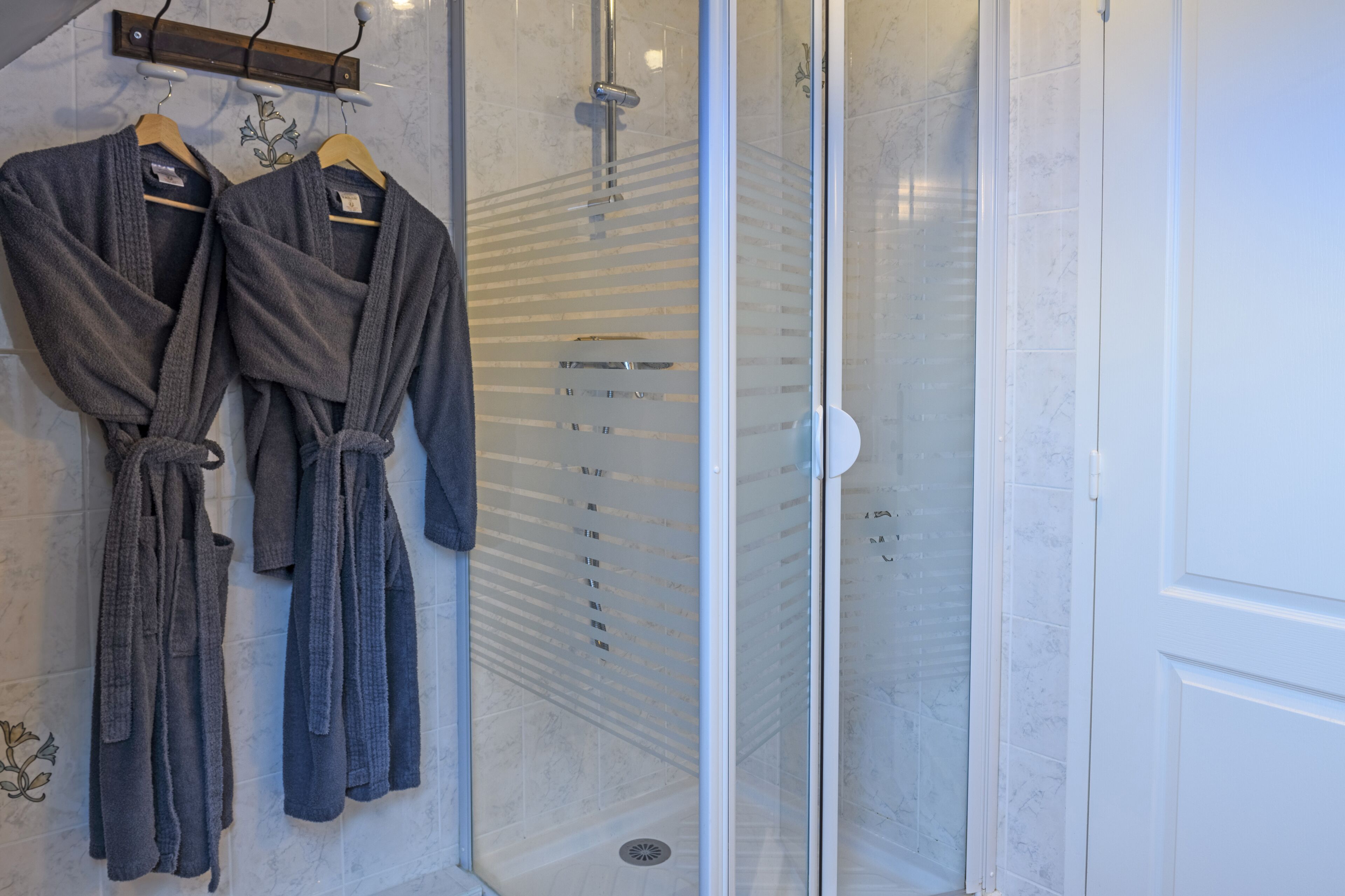 Un baño con dos albornoces grises colgados en un perchero, una cabina de ducha de cristal y una puerta blanca.
