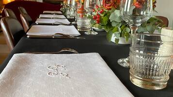 A dining table set with white napkins, silverware, and clear glassware, with a red flower arrangement and a bookshelf in the background.