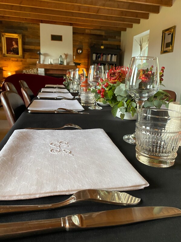 Una mesa de comedor dispuesta con servilletas blancas, cubiertos y copas de cristal transparente, con un arreglo floral rojo y una estantería al fondo.
